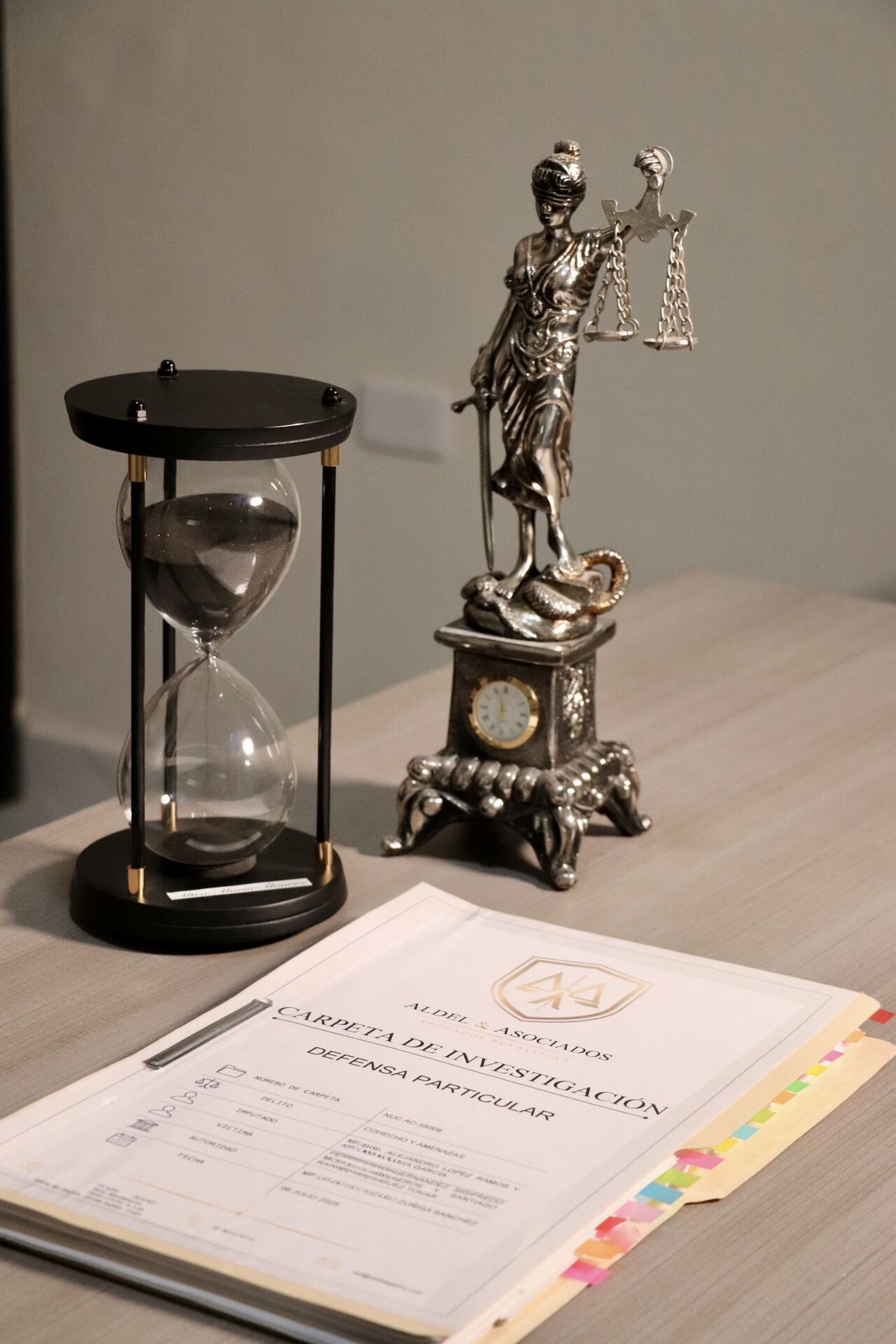 Reloj de arena y balanza de la justicia junto a una carpeta de investigación de ALDEL & Asociados